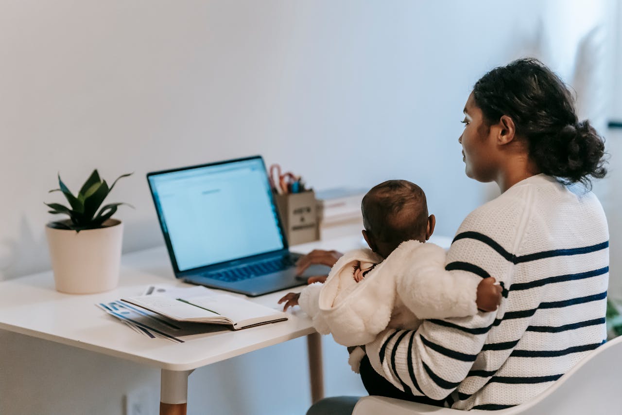 Pensive ethnic woman in casual clothes slitting with baby while browsing netbook in light room in apartment in daytime